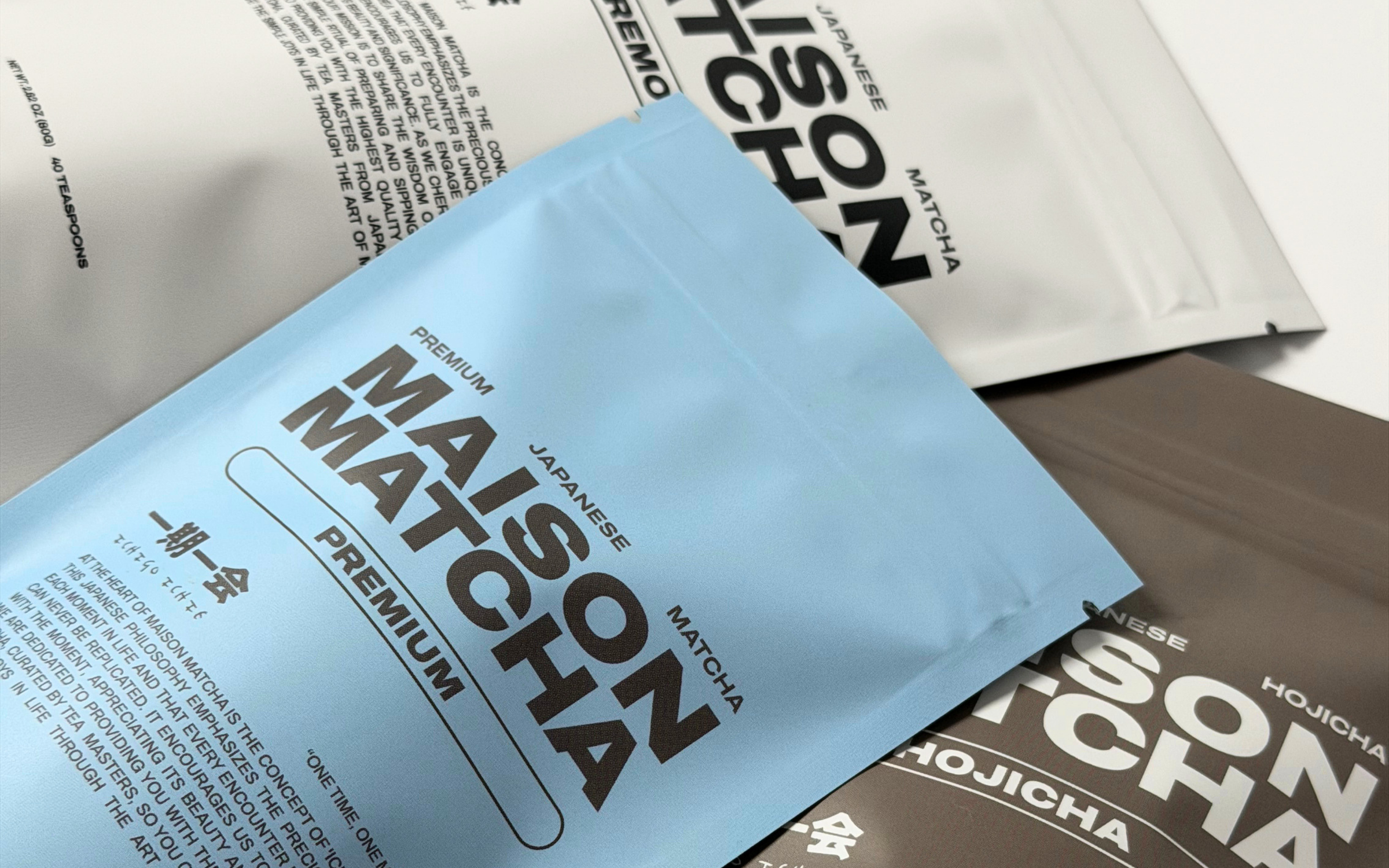 A close-up of a light blue stand-up pouch labeled 'MAISON MATCHA PREMIUM' with black text, layered over white and dark brown pouches on a light background.