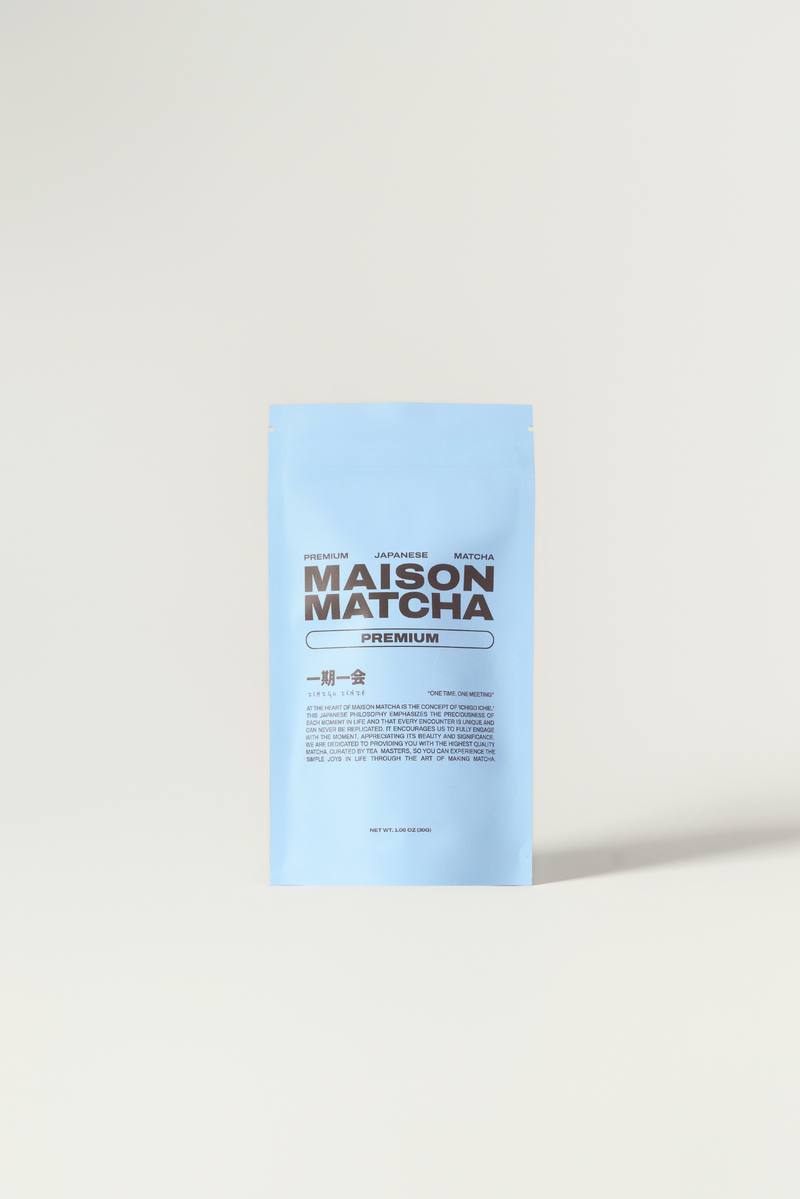 A light blue stand-up pouch of Maison Matcha matcha powder, featuring dark brown text, positioned on a light beige surface against a white background.